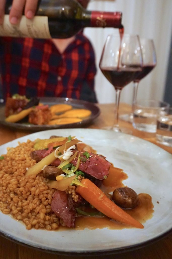 Plate of duck with carrots and barley with au jus and wine being poured in the backgroun