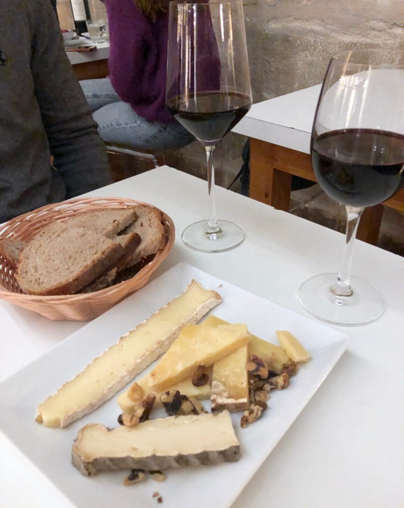 Cheese and nuts on a white plate with a bread basket and two glasses of red wine